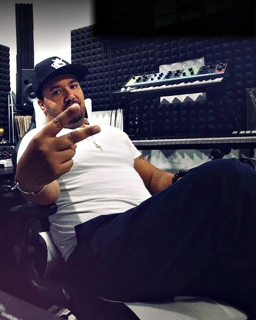 Nick DeAngelis in his studio, leaning back in a chair in a black cap and white T-shirt and holding two fingers up to the camera, with a synthesizer and acoustic foam panels behind him.